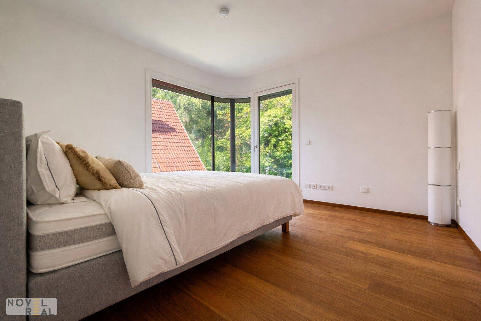 Schlafzimmer mit Holzboden, großen Fenstern und Blick auf die umliegende Natur und Dächer.