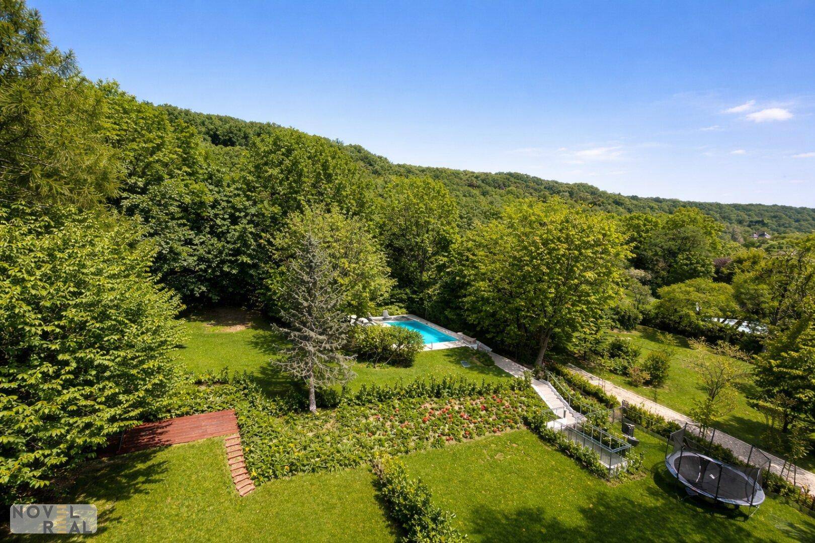 Weitläufige Grünfläche mit Pool, Trampolin und Wegen, umgeben von dichtem Wald und Hügellandschaft.