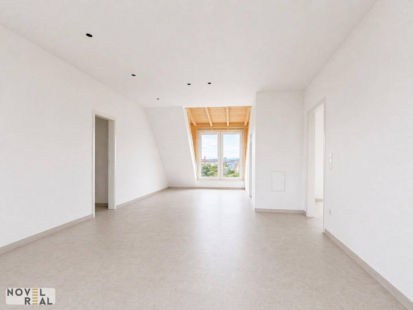 Offener Dachgeschossraum mit schrägen Wänden und einem großen Fenster, das viel Licht hereinlässt.