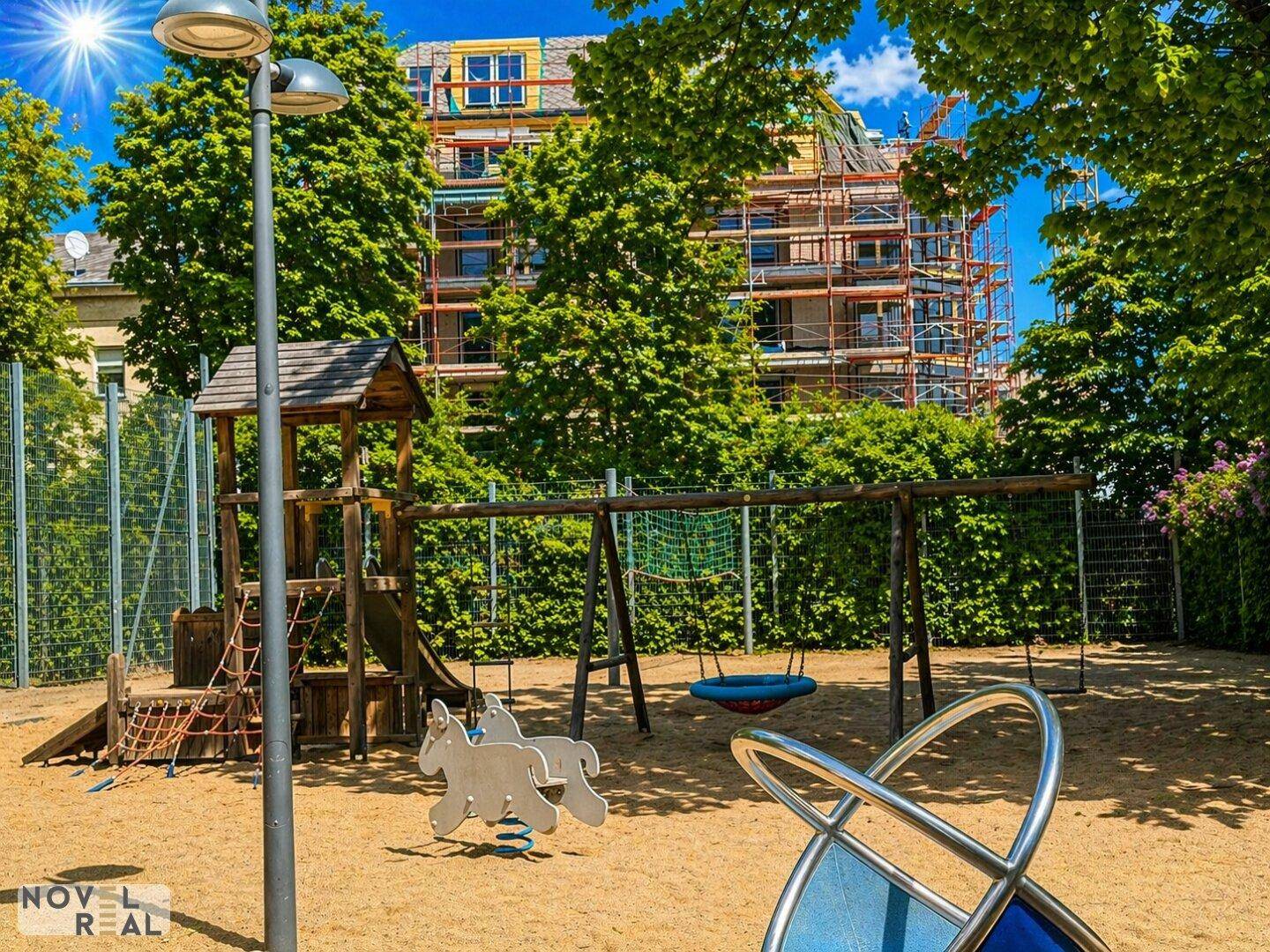 Kinderspielplatz mit Schaukeln und Rutsche, umgeben von Bäumen, im Hintergrund ein Gebäude.