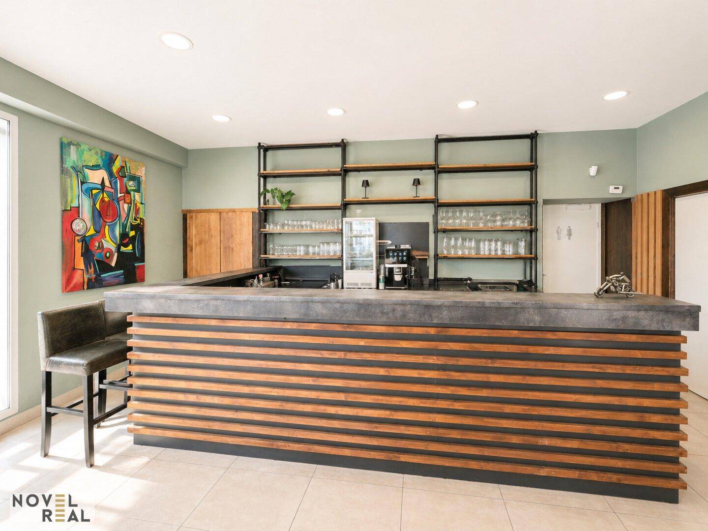Moderne Bar mit einer langen Theke aus Beton und Holzlamellen, dahinter offene Regale für Gläser und Getränke.