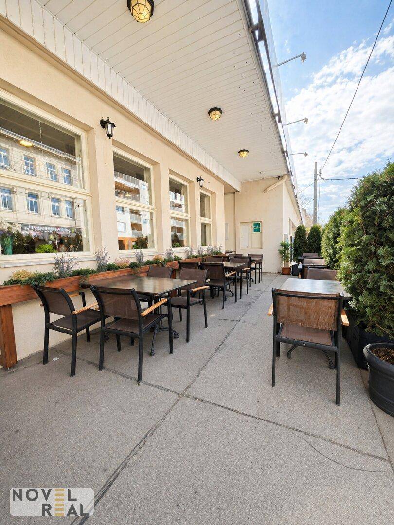 Außenterrasse eines Restaurants mit Tischen und Stühlen, umgeben von grünen Pflanzen und mit Blick auf die Straße.