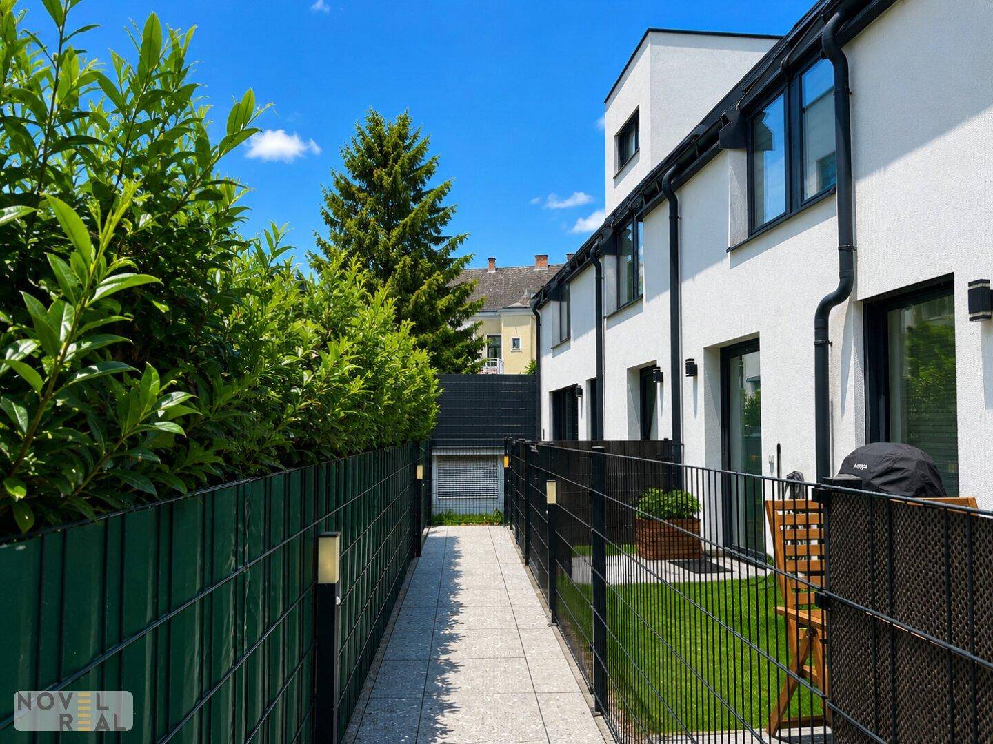 Schmaler Weg zwischen den Häusern, gesäumt von hohen Hecken und einem kleinen Gartenbereich.