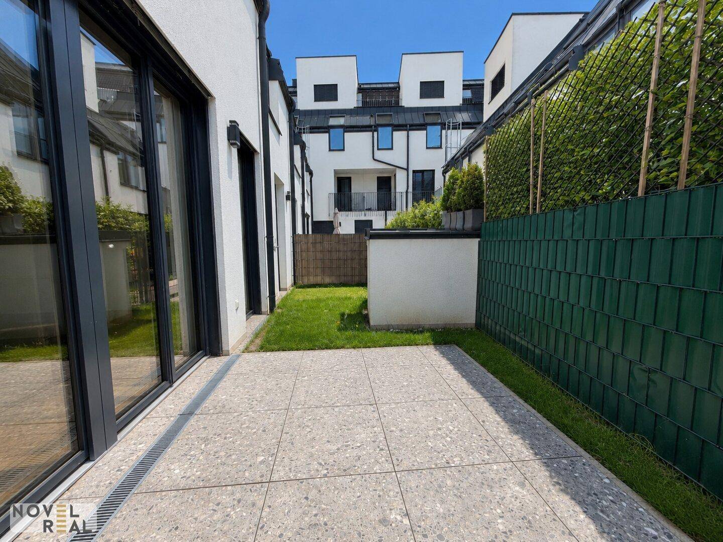 Private Terrasse mit großformatigen Fliesen und angrenzender Rasenfläche, umgeben von Sichtschutz.