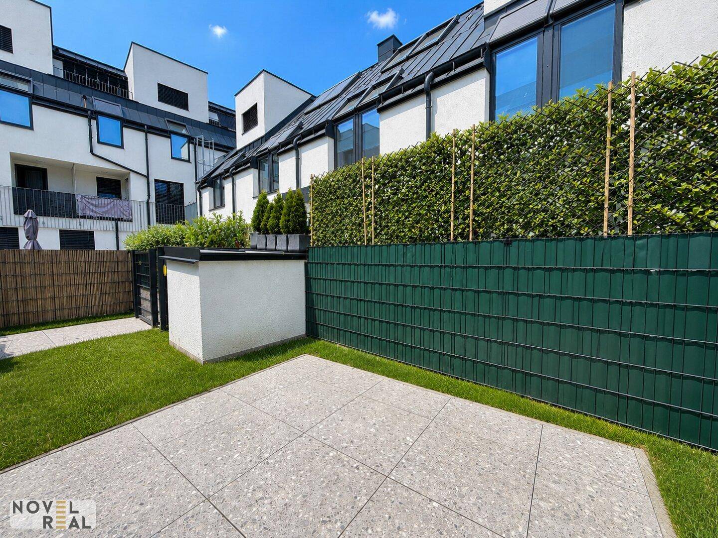 Sonniger Terrassenbereich mit Fliesenboden und gepflegter Rasenfläche, geschützt durch eine hohe Hecke.
