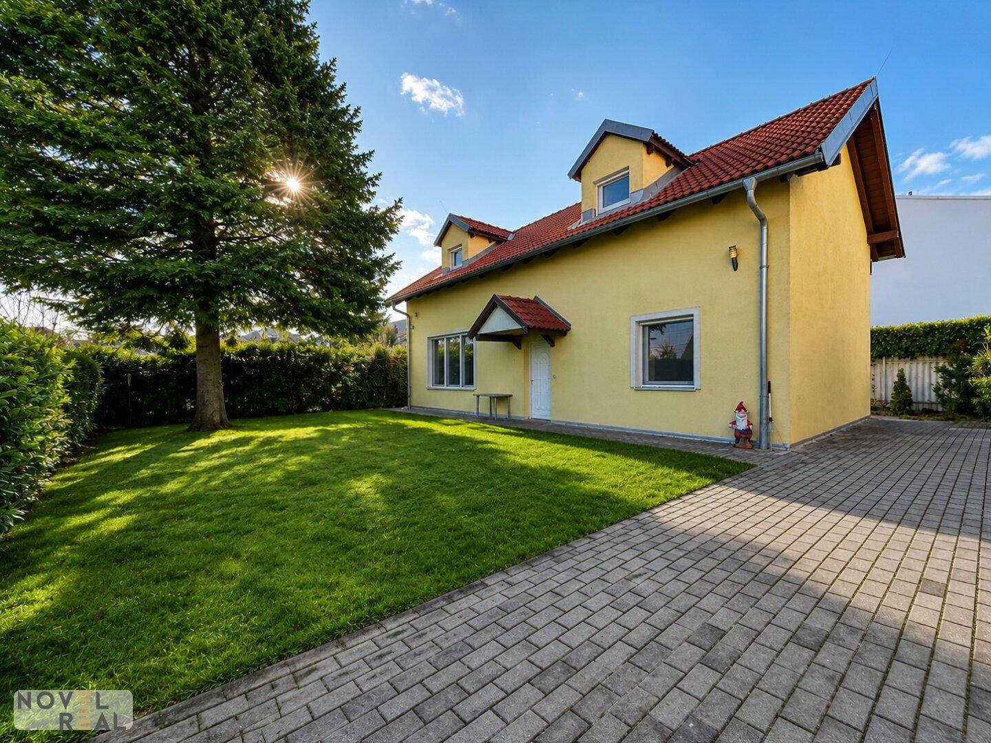 Frontansicht des Hauses mit rotem Ziegeldach, großem Baum und sonnigem Rasenbereich.