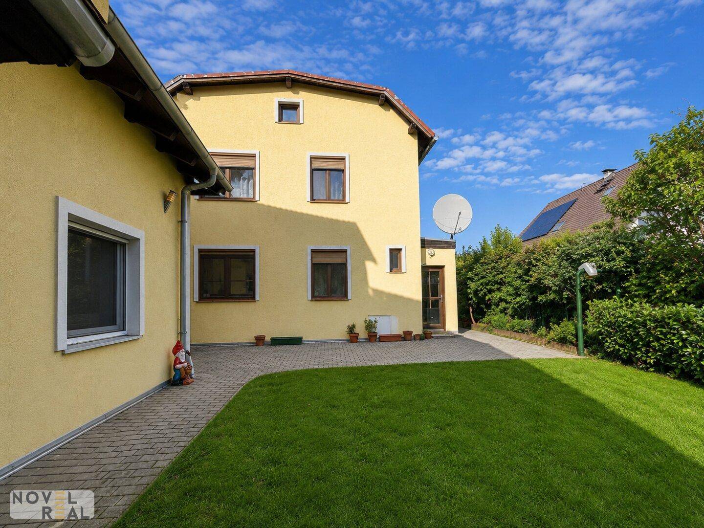 Seitenansicht des Hauses mit Satellitenschüssel, gepflastertem Weg und grünem Rasen unter blauem Himmel.