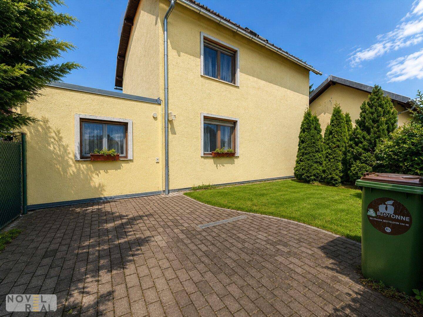 Rückansicht des Hauses mit gepflasterter Einfahrt, Rasenfläche und Mülltonne im Gartenbereich.