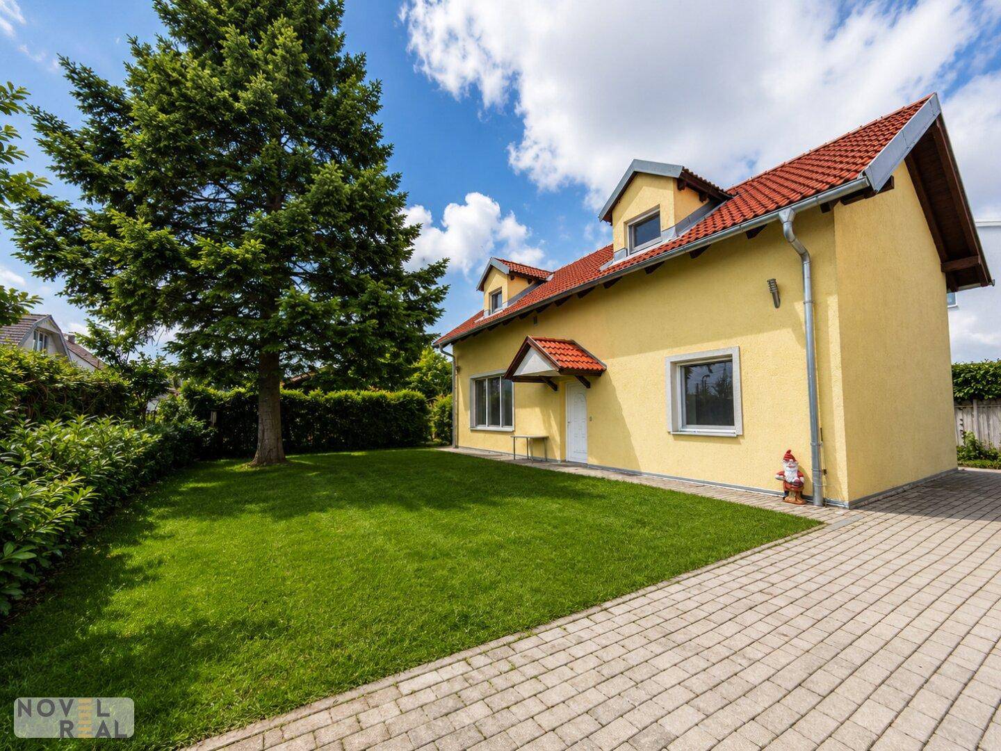 Seitenansicht des gelben Hauses mit rotem Dach, großem Baum und gepflegtem Rasen unter blauem Himmel.