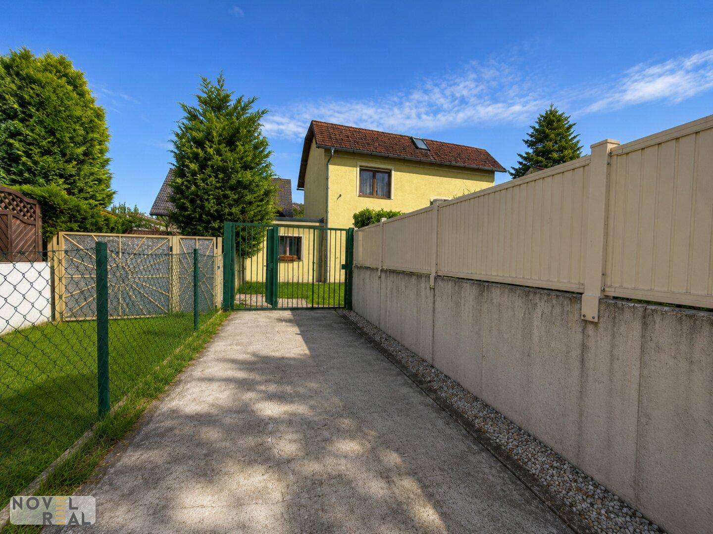 Zufahrtsweg zum Haus mit grünem Zaun und Blick auf das gelbe Gebäude im Hintergrund.