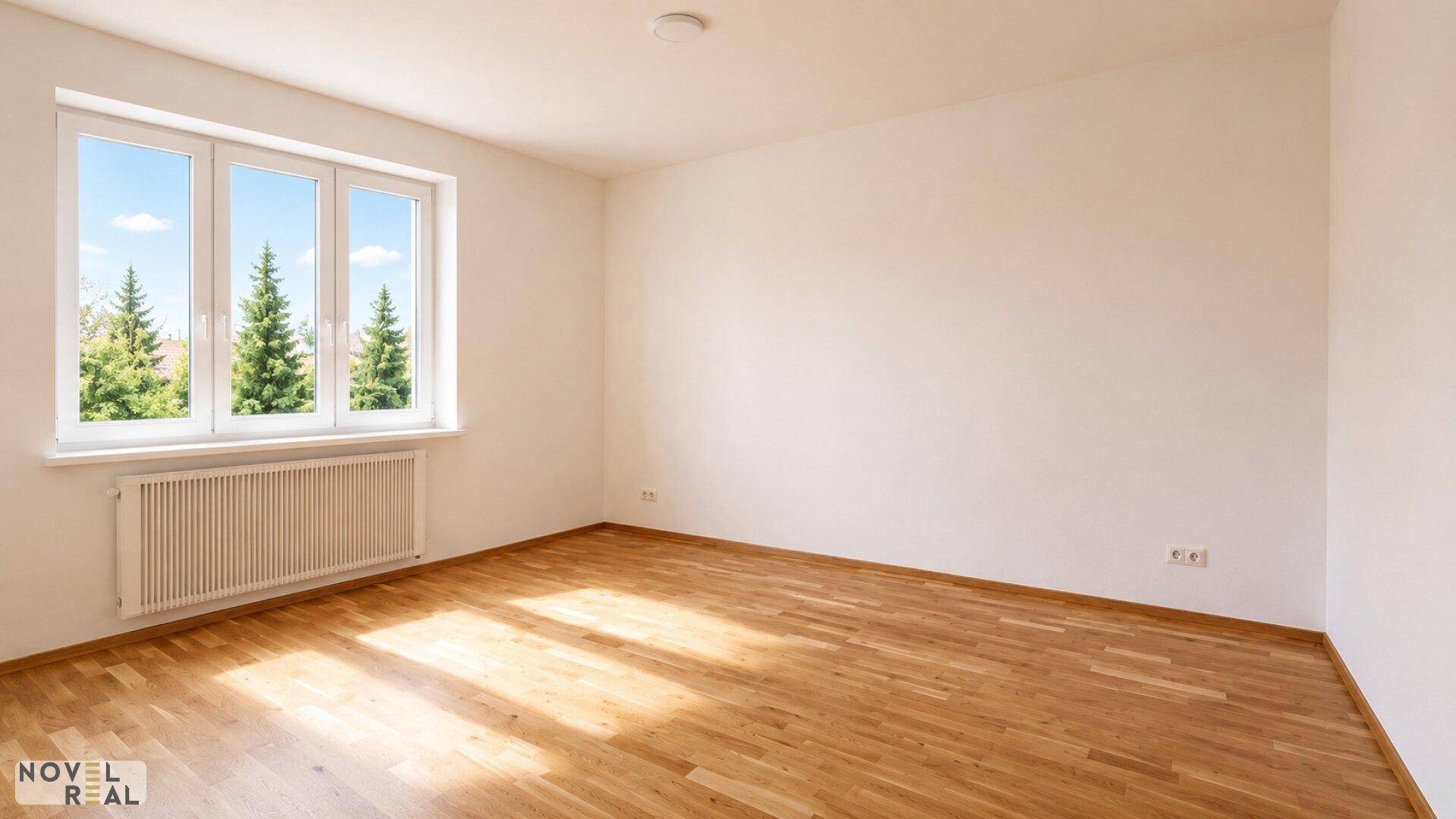 Helles, leeres Zimmer mit Holzboden und großen Fenstern mit Blick auf Bäume.