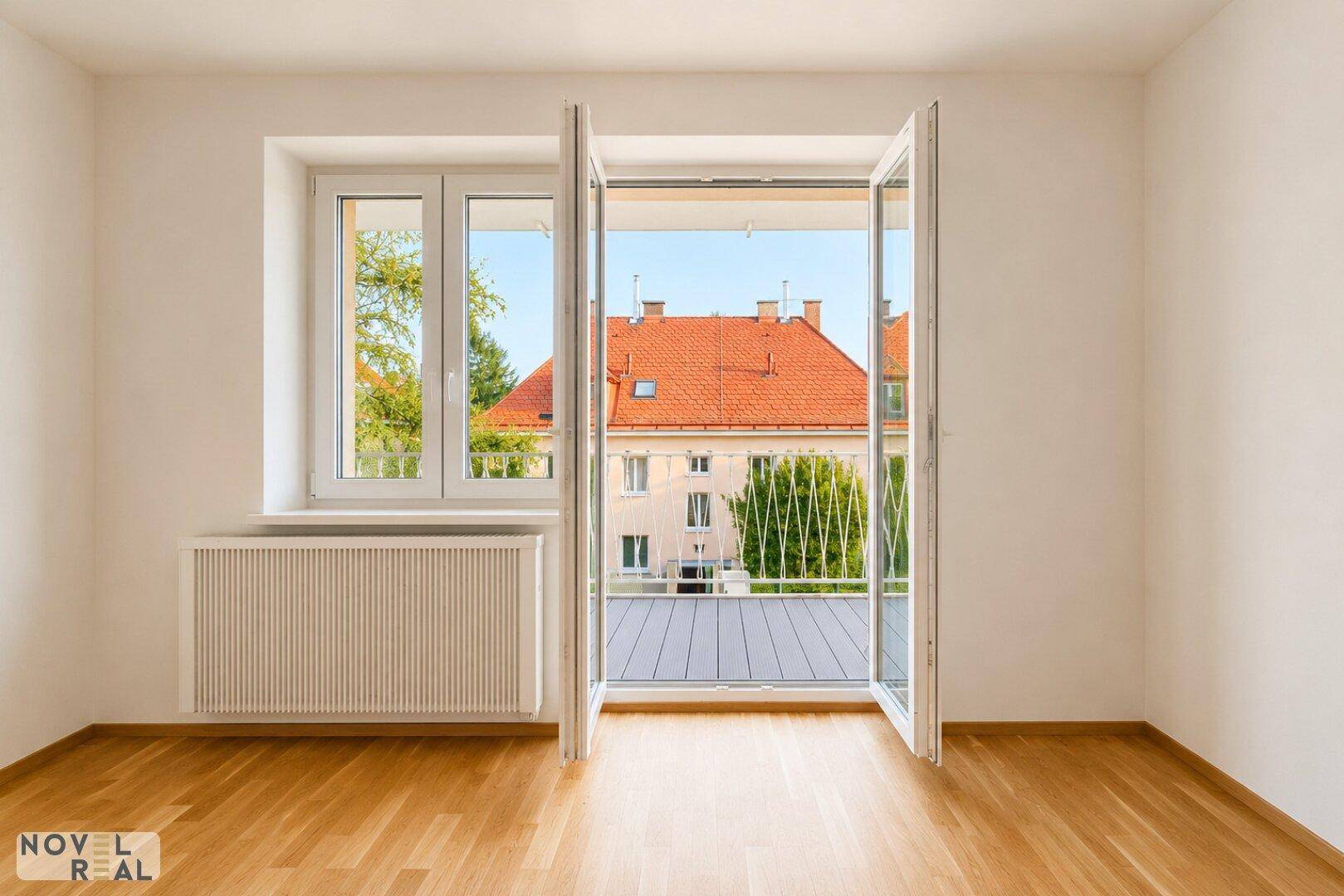 Heller Raum mit Holzboden und geöffneten Fenstern, die den Blick auf die Nachbarschaft freigeben.