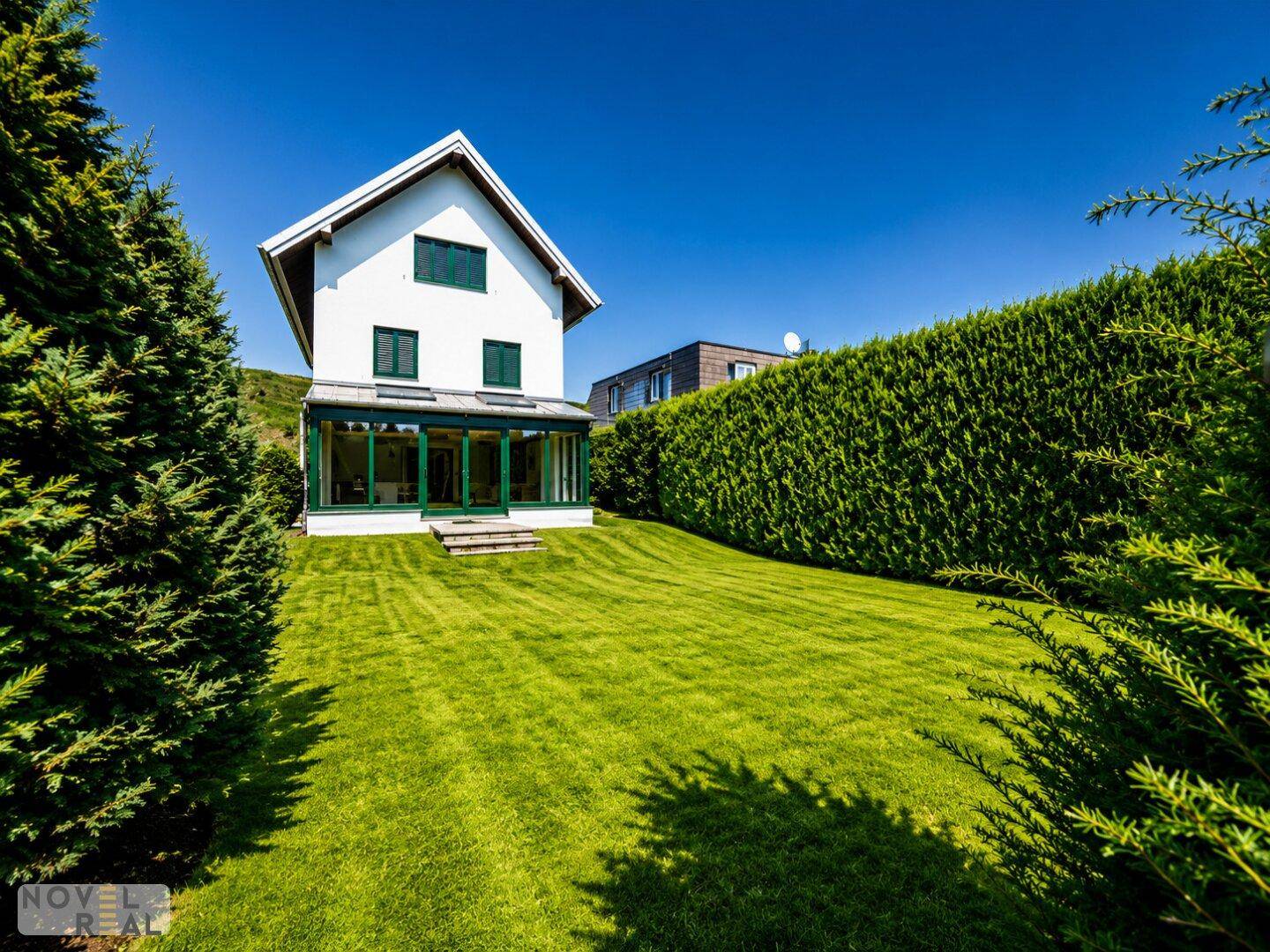 Weitläufiger Garten mit grüner Hecke und Blick auf das Haus mit großem Wintergarten.