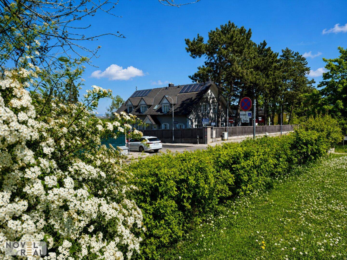 Traditionelles Haus mit Solaranlage auf dem Dach, umgeben von üppigem Grün und blühenden Sträuchern an einem sonnigen Tag.