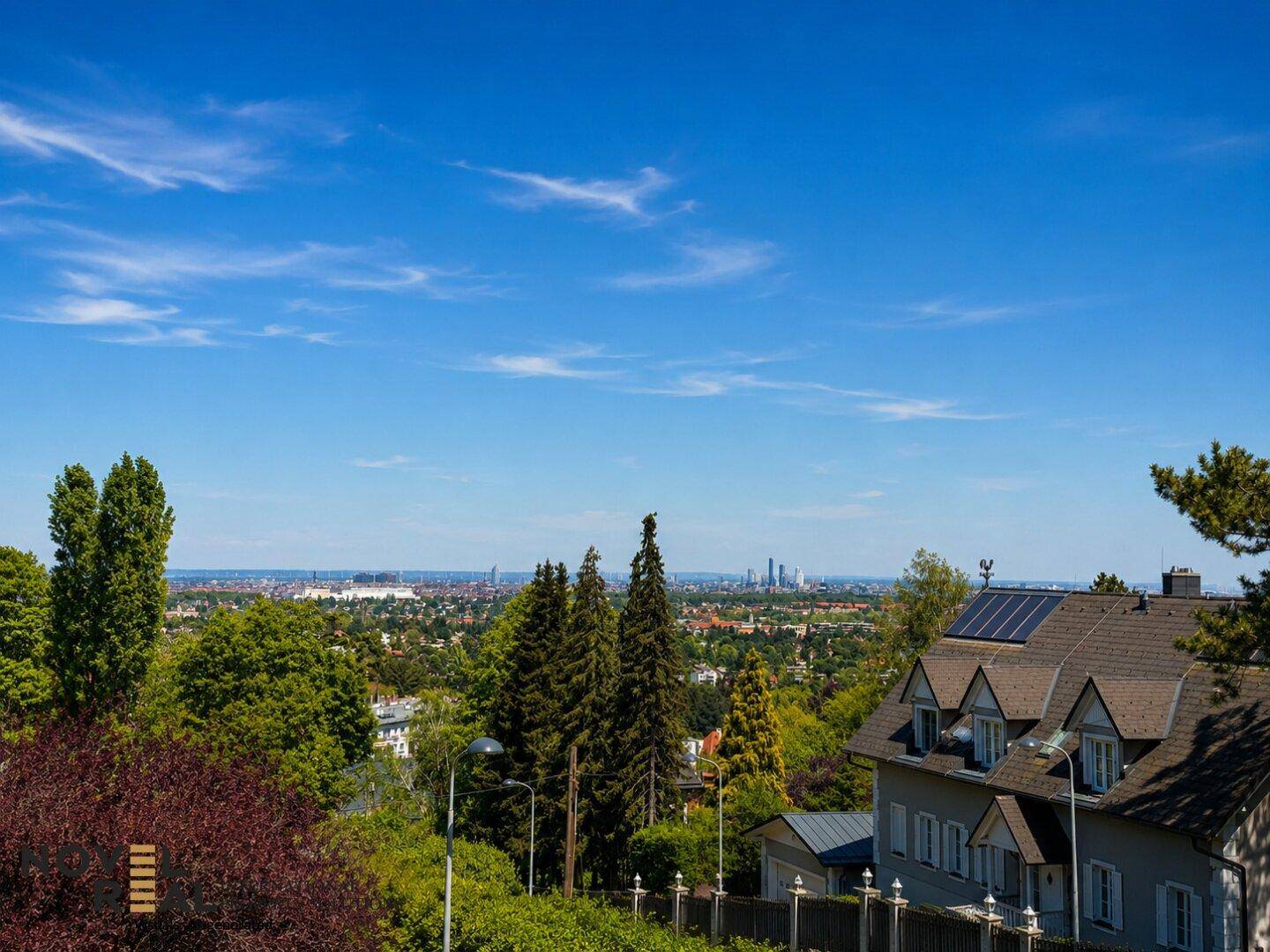 Panoramablick über eine Stadtlandschaft von einem grünen Hügel mit Wohnhäusern und Bäumen.