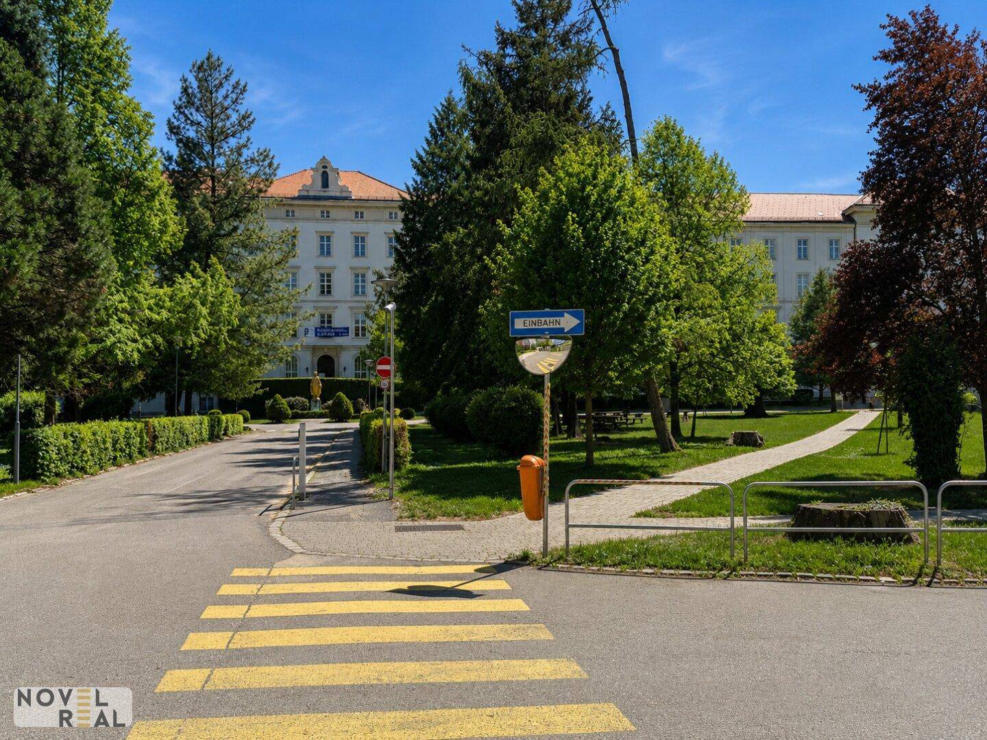 Zufahrt zu einem großen Gebäude mit gepflegten Grünflächen und Bäumen unter blauem Himmel.