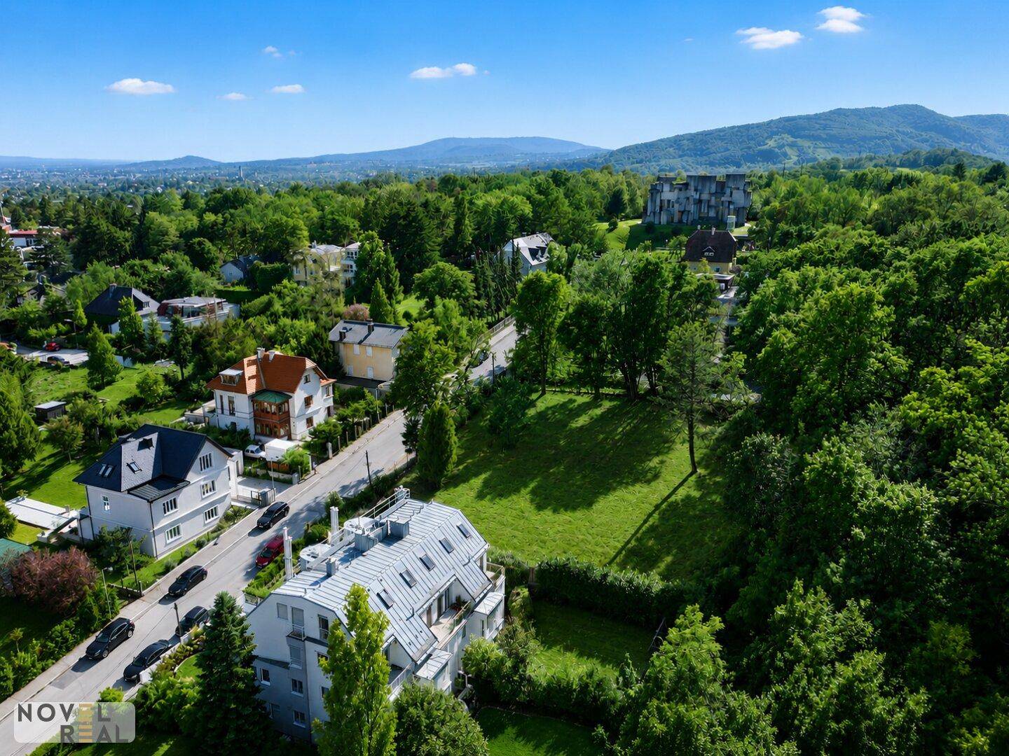 Luftaufnahme einer Wohnstraße mit Häusern, Grünflächen und Bergblick in der Ferne.
