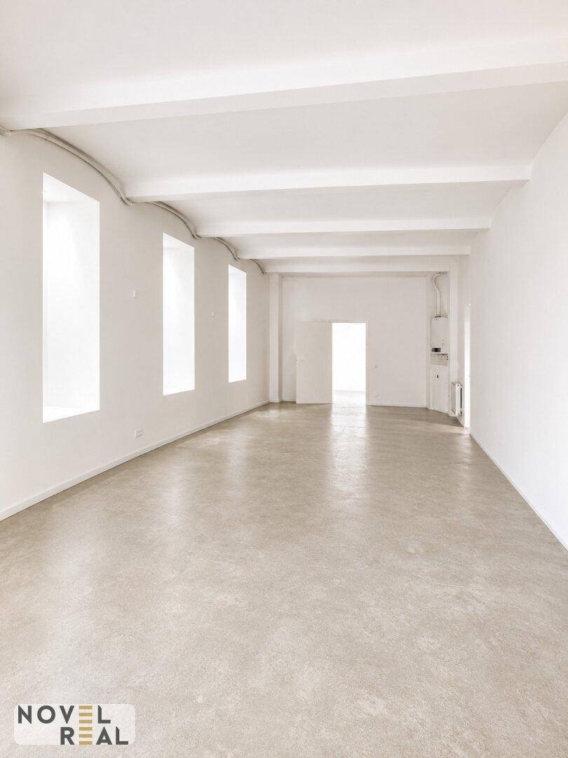 Weitläufiger, heller Raum mit Betonboden, hohen Fenstern und sichtbaren Deckenbalken, ideal für flexible Nutzung.