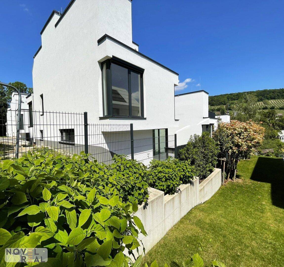 Moderne weiße Architektur mit großen Fenstern, umgeben von grünen Büschen und einem Weinberg im Hintergrund.