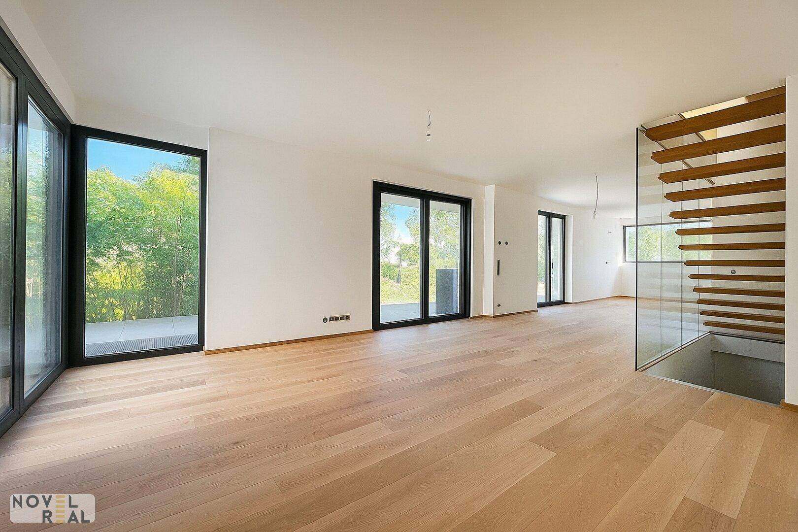 Geräumiger Wohnbereich mit hellem Holzfußboden, großen Fenstern und offener Treppe.