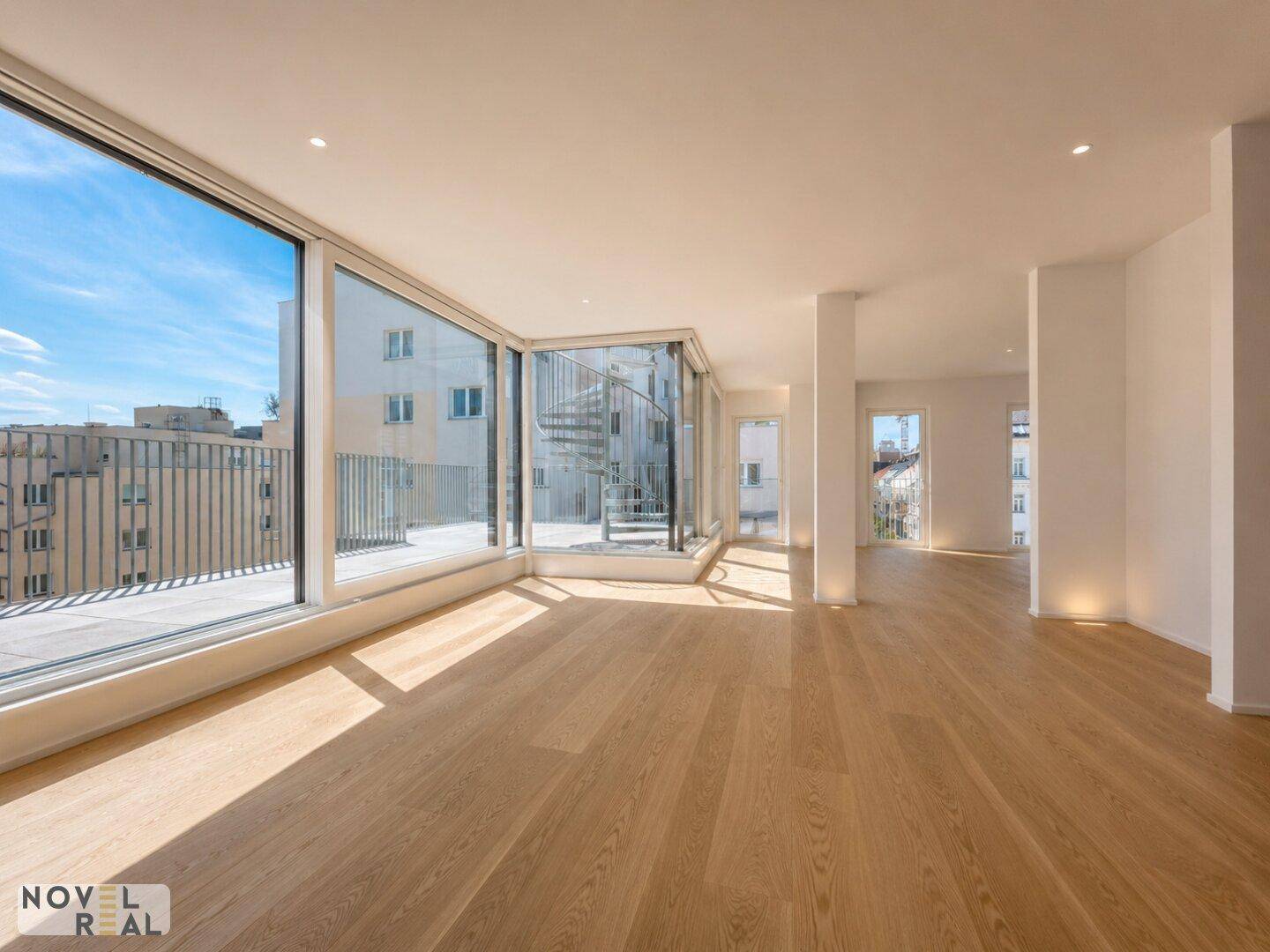 Heller, leerer Wohnraum mit großen Fenstern, Holzfußboden und Blick auf eine Wendeltreppe.