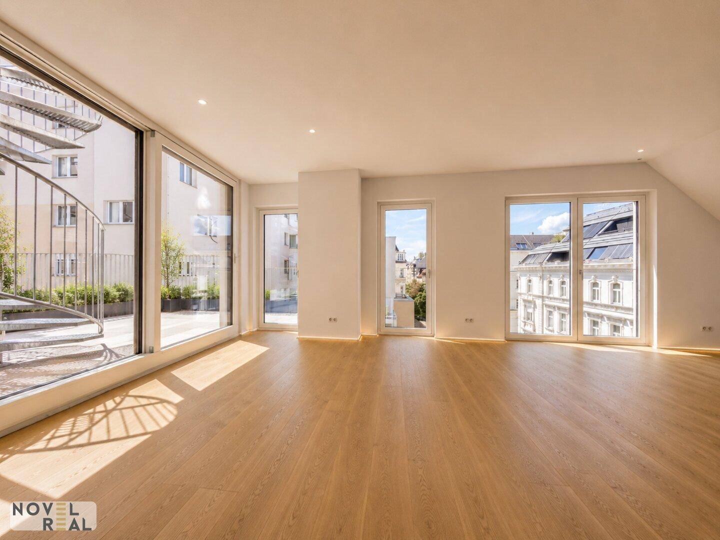 Geräumiger Wohnraum mit Holzfußboden, großen Fenstern und Zugang zu einem Außenbereich.