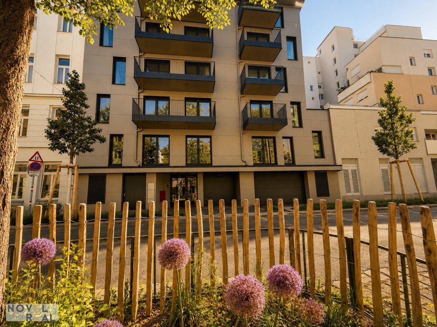 Ansicht eines modernen Wohngebäudes mit Balkonen, umgeben von Blumen und einem Holzzaun.