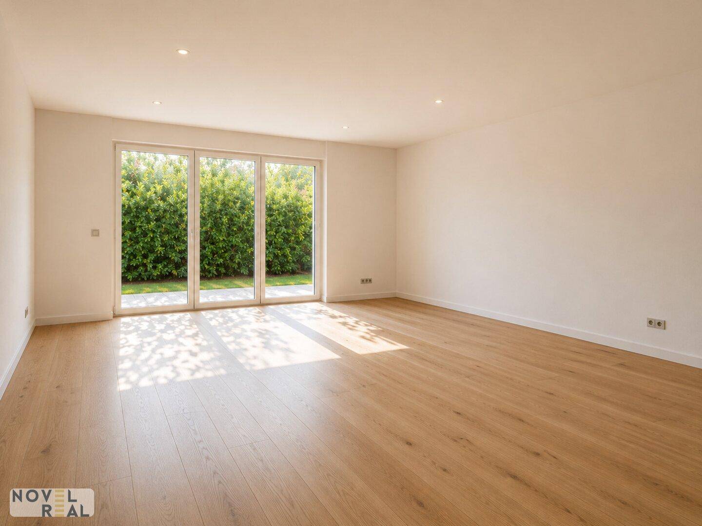 Heller, leerer Raum mit Holzboden und großen Fenstern mit Blick auf eine grüne Hecke.