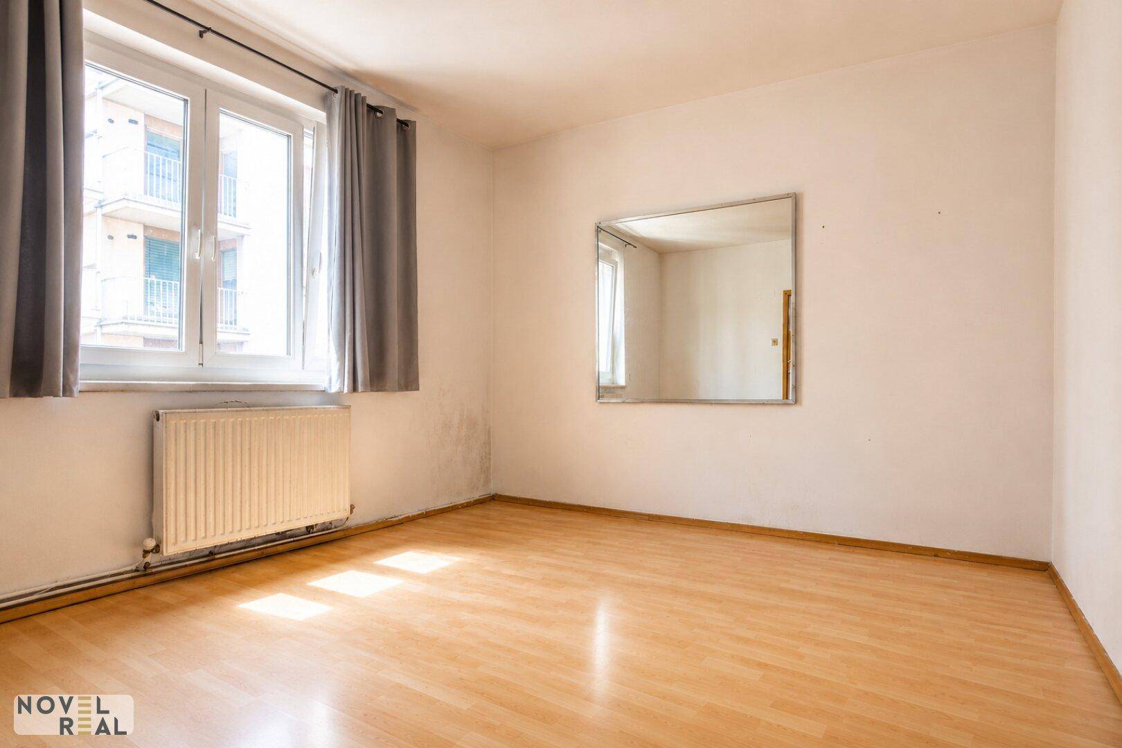 Leeres Schlafzimmer mit hellem Holzfußboden, großem Fenster und einem Wandspiegel.