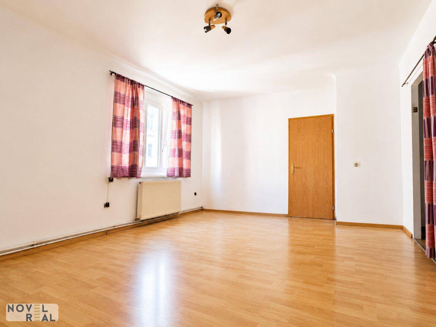 Helles, leeres Zimmer mit Holzfußboden, Fenster und Heizkörper, bereit zur individuellen Gestaltung.