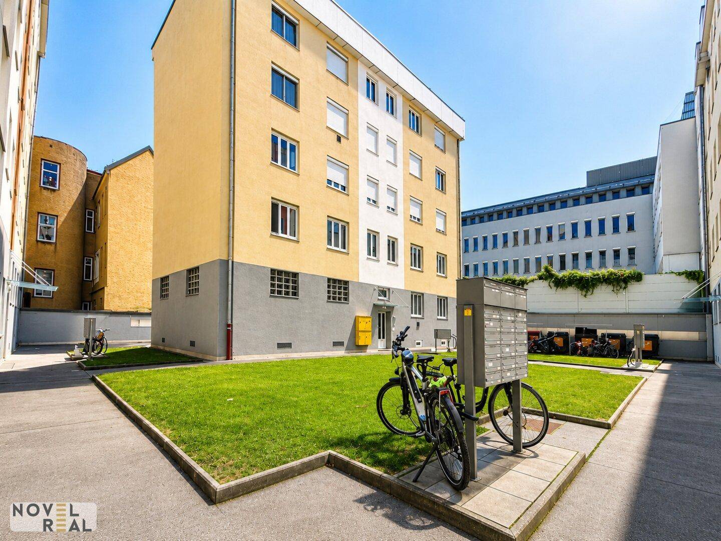 Wohngebäude mit gelber Fassade, Grünfläche, Briefkästen und Fahrradständern im Außenbereich.
