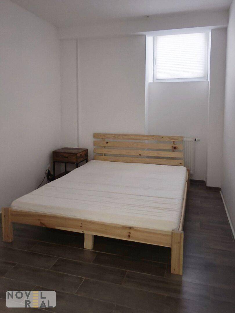 Schlafzimmer mit einfachem Holzbettrahmen, weißer Matratze und Fliesenboden in Holzoptik.