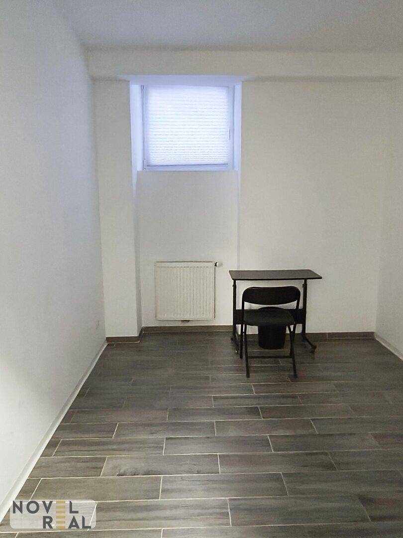 Heller, leerer Raum mit Fliesenboden in Holzoptik, weißen Wänden und einem Fenster mit Jalousien.
