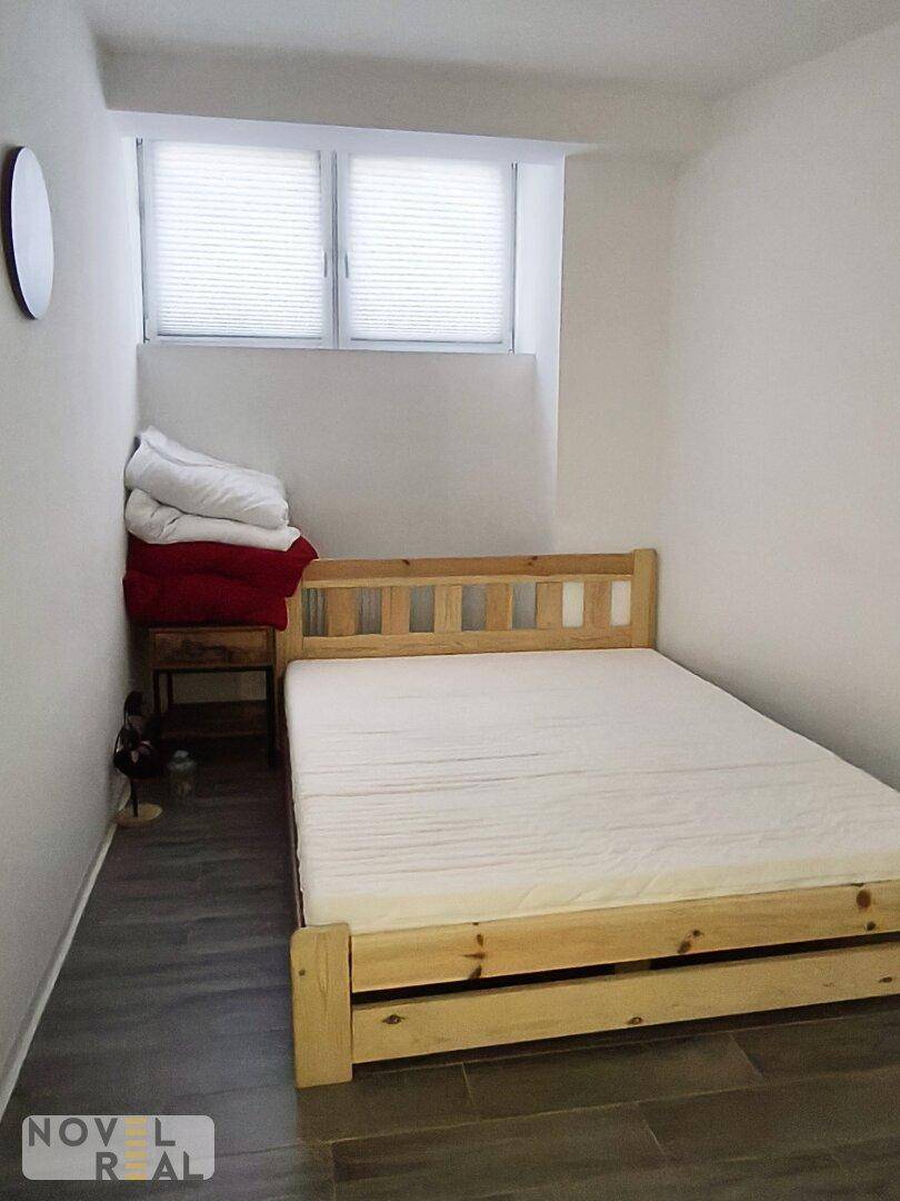 Schlafzimmer mit einfachem Holzbettrahmen, weißer Matratze und Fliesenboden in Holzoptik.