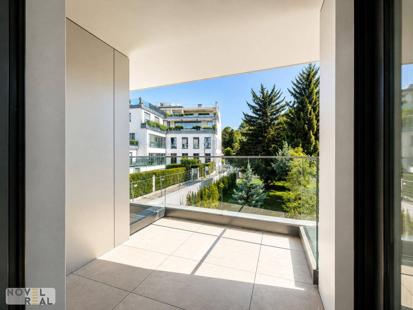 Geräumiger Balkon mit Fliesenboden und Glasgeländer, bietet einen weiten Blick ins Grüne.