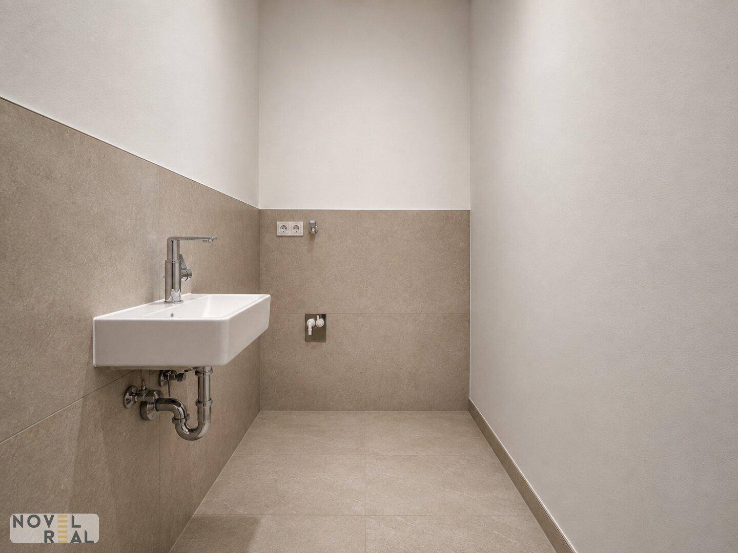 Modernes Gäste-WC mit beigefarbenen Fliesen, kleinem Waschbecken und Platz für eine Waschmaschine.