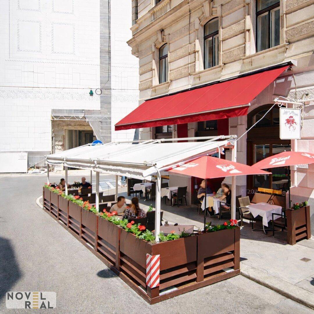 Belebte Außenterrasse eines Restaurants mit Sonnenschirmen, Markisen und Blumenkästen an der Straße.