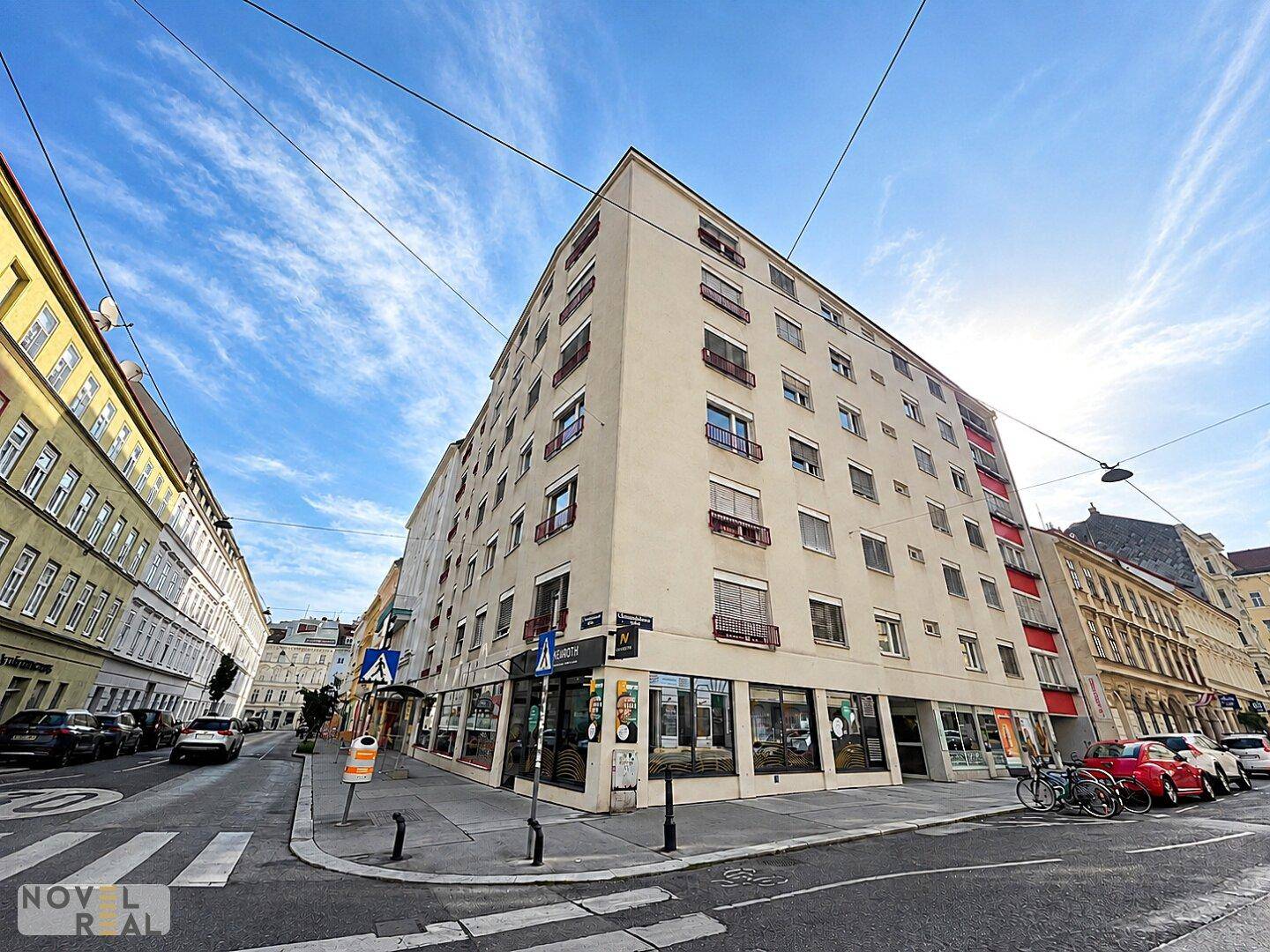 Eckgebäude in einer städtischen Umgebung mit Geschäften im Erdgeschoss und vielen Fenstern.