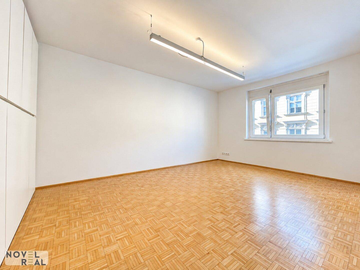 Geräumiger, heller Raum mit Mosaikparkettboden und großen Fenstern, ideal als Wohn- oder Schlafzimmer.