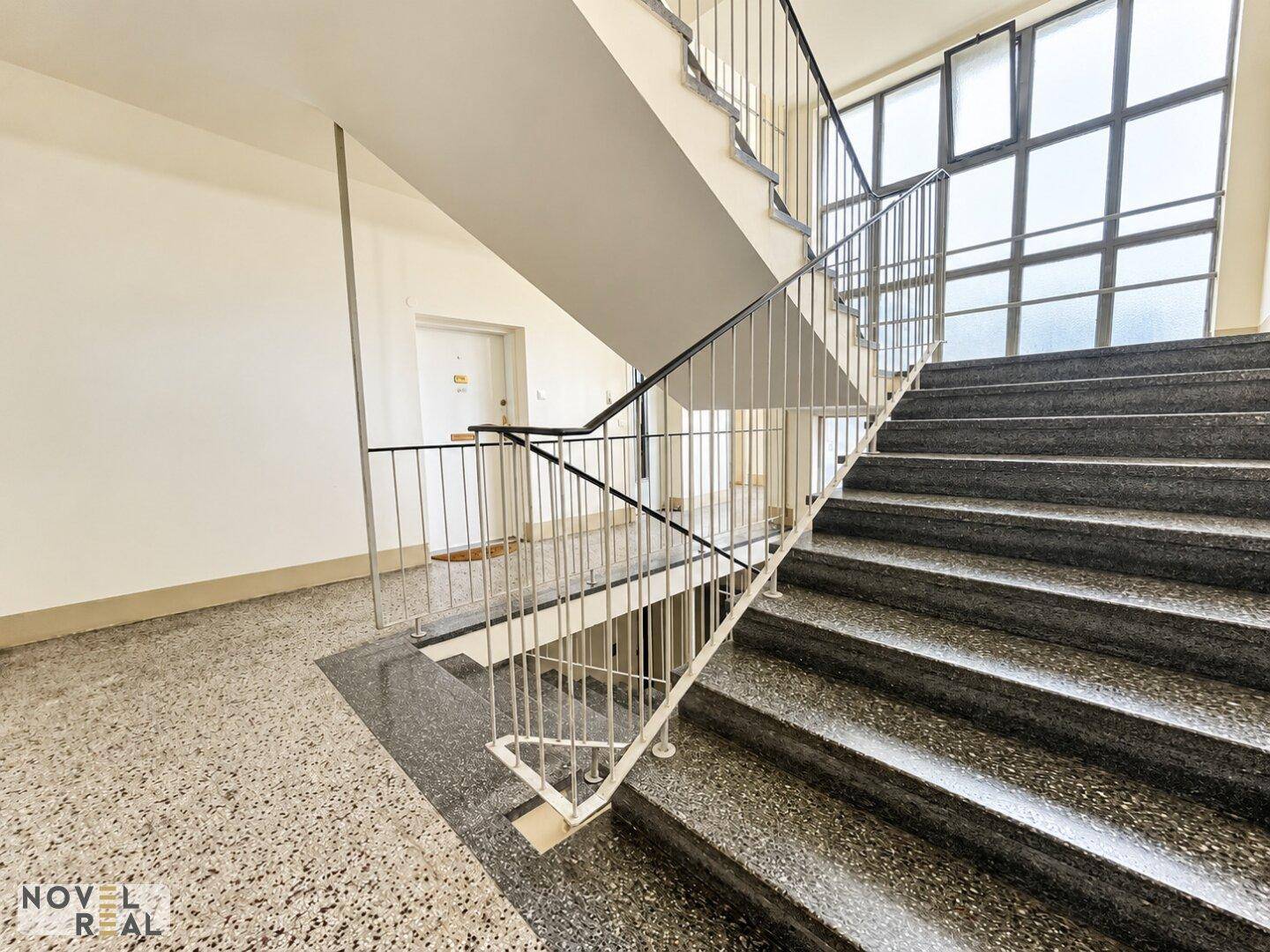 Treppenhaus mit Terrazzoboden und Metallgeländer, das zu den oberen Etagen führt.