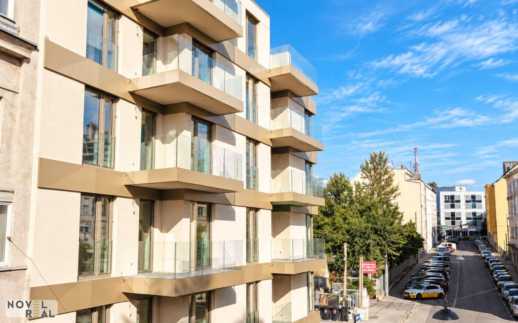 Moderne Wohnhausfassade mit zahlreichen Balkonen und großen Fenstern, umgeben von städtischer Umgebung.