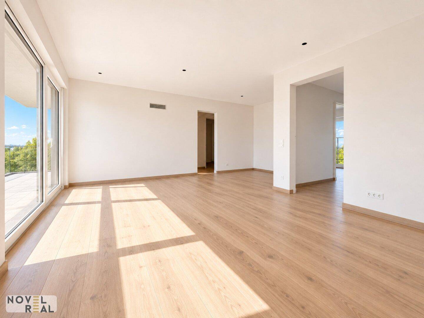 Geräumiges, leeres Wohnzimmer mit hellem Holzboden und großen Fenstern, die viel Sonnenlicht hereinlassen.