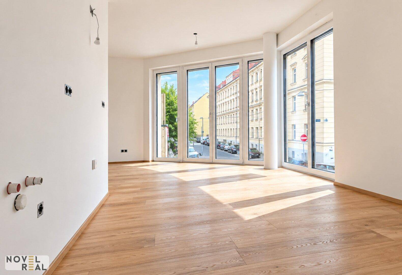 Heller Raum mit großen Fenstern, Holzboden und Anschlüssen für eine Küche.