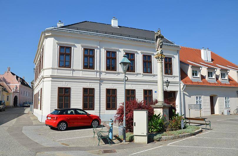 Ansicht eines weißen historischen Gebäudes mit braunen Fenstern und einem roten Auto auf dem Vorplatz.