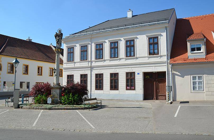 Historisches weißes Gebäude mit braunen Fenstern und einem Denkmal auf dem gepflasterten Vorplatz.