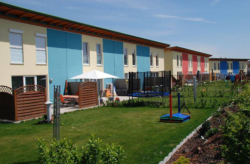 Sonnige Gärten hinter modernen Reihenhäusern mit Trampolin und Sitzbereichen für Familien.