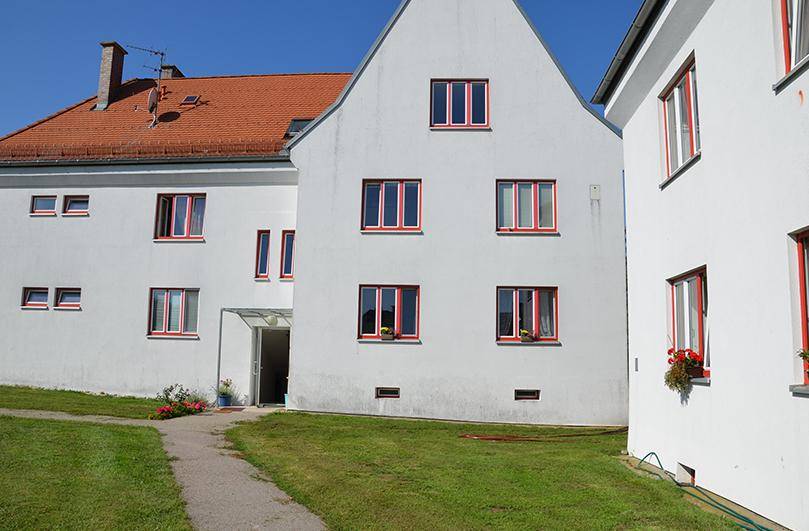 Vorderansicht eines weißen Gebäudes mit roten Fensterrahmen und einem kleinen Eingangsbereich.