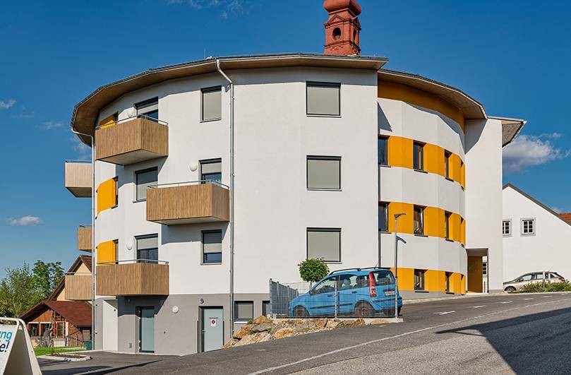 Rundes Wohngebäude mit orangefarbenen Fassadenakzenten, Balkonen und einem roten Kirchturm im Hintergrund.