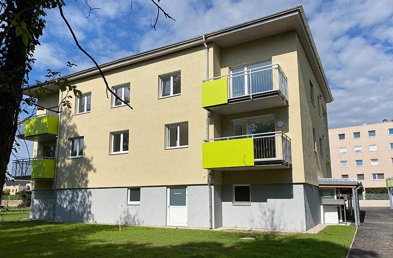 Seitenansicht eines modernen Wohngebäudes mit hellgelber Fassade, grünen Balkonen und einem gepflegten Rasenbereich.