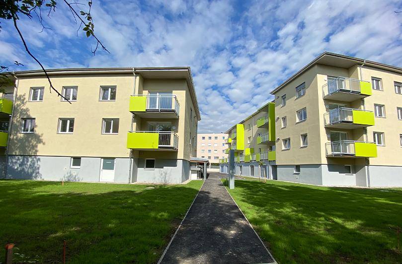 Zwei moderne Wohngebäude mit hellgelben Fassaden und grünen Balkonen, verbunden durch einen Gehweg.